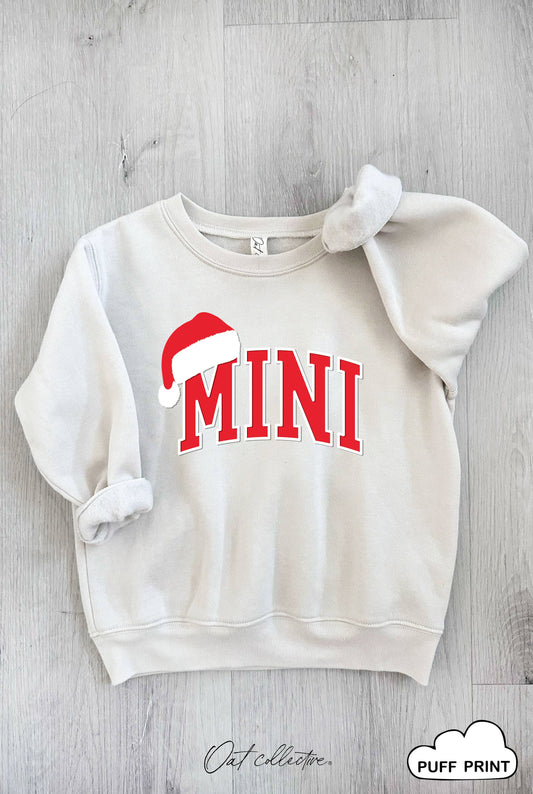 White sweatshirt with 'MINI' in red and a Santa hat graphic on a gray wooden surface.